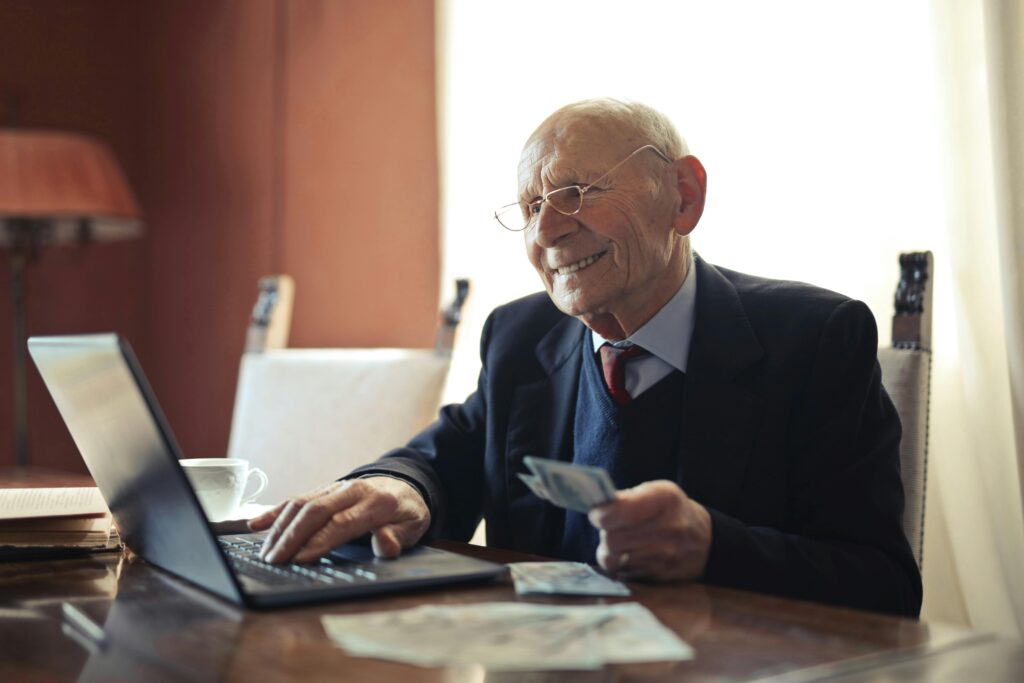 Senior man in formal attire using a laptop and holding money, symbolizing financial success.