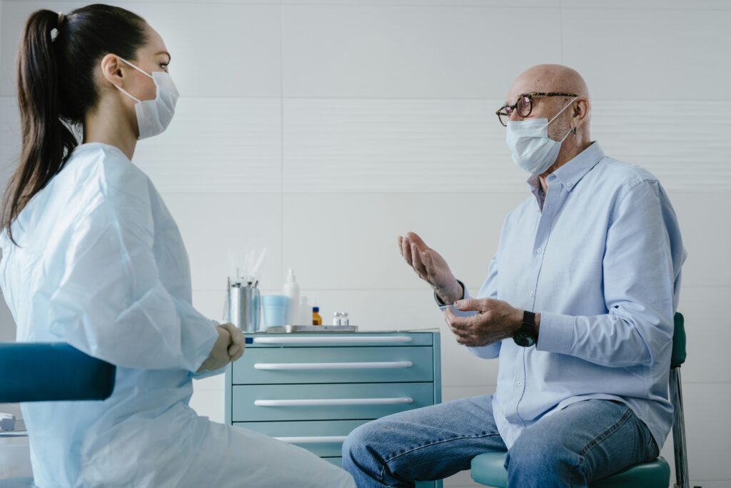 A senior adult patient discusses health concerns with a masked medical professional in a clinic setting.