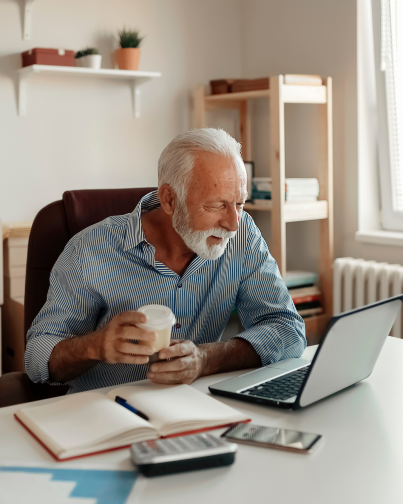 Senior at desk working on laptop with coffee in hand